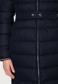 Manteau en duvet navy avec un motif matelassé horizontal, fermeture éclair métallique et taille élastique avec deux pressions. Présente des poignets intérieurs contrastants.