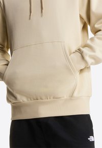 Beige hoodie gemaakt van zachte stof, met een capuchon met trekkoord, een kangoeroezak en geribde zoom en manchetten, gecombineerd met een zwarte broek.