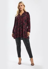 Donna bionda che indossa una tunica button-up a maniche lunghe di colore bordeaux e nero a motivi, abbinata a leggings in pelle nera e tacchi neri.