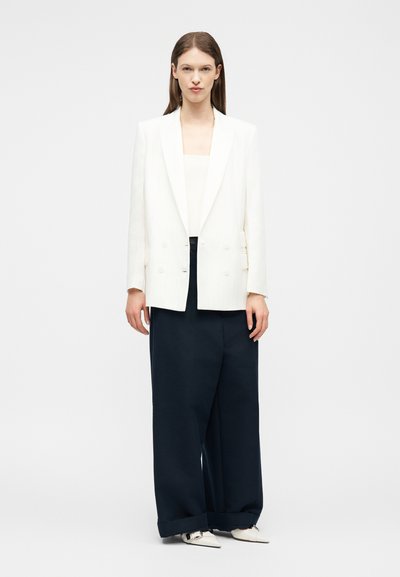 Jeune femme portant un blazer blanc à double boutonnage, un haut blanc, un pantalon large noir et des chaussures blanches pointues, debout devant un fond blanc.