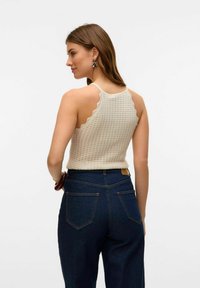 Top in crochet di colore beige con bordi a scallop, abbinato a jeans di denim blu a vita alta con tasche posteriori. I capelli del modello sono sciolti.