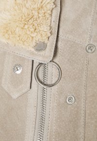 Beige suède jas close-up met zilveren rits, ronde trekker, drukknopen, klepzak en crème kleurige schapenvachtkraag detail.
