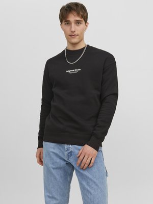 Sweatshirt noir avec col rond, poignets côtelés et un graphique blanc "Original Studio" sur la poitrine, associé à un jean bleu clair.