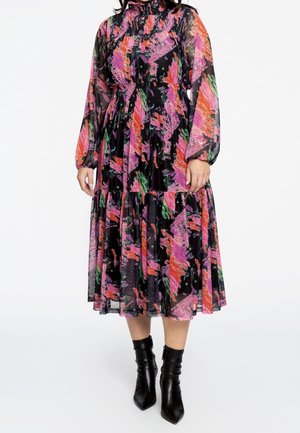 Kobieta ubrana w midi sukienkę z długim rękawem i wysokim dekoltem, ozdobioną kolorowym, abstrakcyjnym wzorem w odcieniach różu, pomarańczy, zieleni i czerni, zestawioną z czarnymi botkami za kostkę.