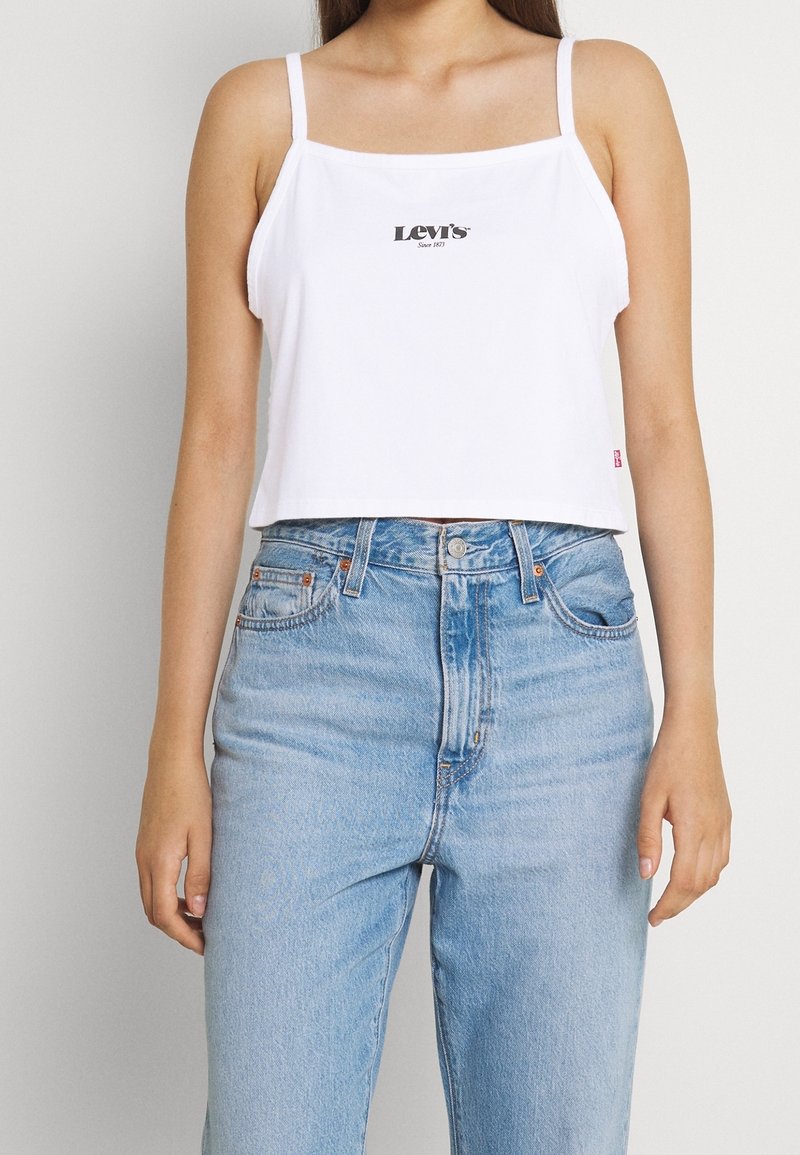 Vit crop top med spaghettistölkar och en svart "Levi's" logga i mitten, kombinerad med ljusblå högmidjade jeans.