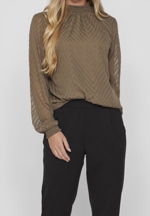 Femme portant un chemisier transparent vert olive texturé à manches longues avec poignets à volants et un pantalon noir taille haute, debout devant un fond uni.