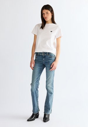 Fiatal nő áll, fehér rövid ujjú pólót, kék szűk farmert és fekete hegyes orrú bokacsizmát viselve, fehér háttér előtt.