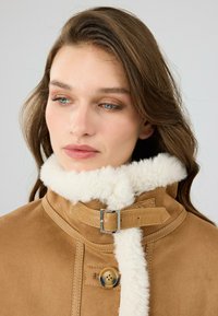 Tan suède jack met een witte nep bont kraag, zilveren hardware en een knoopsluiting aan de voorkant. Zachte textuur, op maat ontworpen.