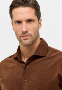 Bruin corduroy shirt met een brede kraag, voorzien van zwarte knopen en een zachte textuur, getoond op een model tegen een lichte achtergrond.