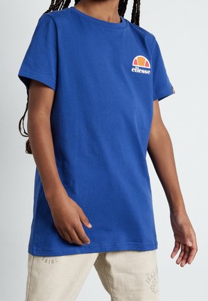 Person trägt ein blaues ellesse T-Shirt und hellbeige Hosen, steht mit entspannten Armen vor einem einfarbigen hellen Hintergrund.