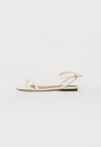 Flat sandals in cream leather with thin cross straps, ankle strap with buckle, minimalistic design, and a contrasting black sole.