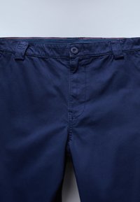 Pantalones azul marino con cinturilla abotonada, trabillas para cinturón y costuras visibles sobre un fondo claro.