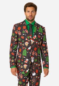 Melns žakets ar svētku Ziemassvētku rakstiem, kuros redzami koki, dāvanas un piparkūku cilvēciņi; kombinēts ar spilgti zaļu kreklu un kaklasaiti.