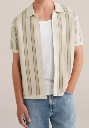 Homme portant une chemise en maille beige à manches courtes avec des rayures verticales, par-dessus un t-shirt blanc et un jean bleu clair, une main dans la poche.