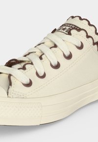 Ανοιχτόχρωνο καμβαδένιο αθλητικό παπούτσι με καφέ κυματιστή μπορντούρα, καφέ μεταλλικά οπές, λευκά κορδόνια και καουτσούκ σόλα.