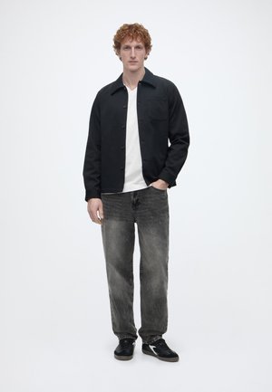 Mladić s kovrčavom kosom u crnoj zakopčanoj jakni, bijeloj majici, izblijedjelim crnim trapericama i crnim tenisicama, stoji s jednom rukom u džepu.