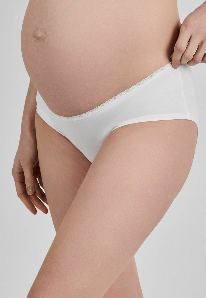 Schwangere Person trägt weiße Umstandsunterwäsche, Hand zieht den Bund sanft vom Bauch weg.