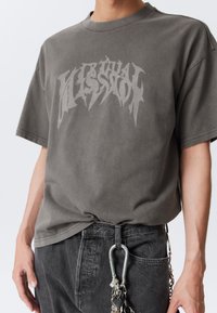 Harmaa ylipitkä t-paita, jossa on kontrastivärinen graafinen painatus. Lyhyet hihat, pyöreä pääntie, pehmeä materiaali. Yhdistetty tummiin farkkuihin ja metallisiin yksityiskohtiin.