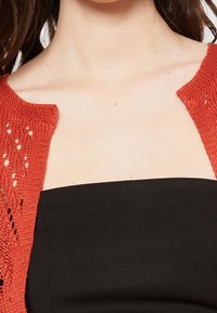 Cardigan aperto lavorato a maglia di colore ruggine, con un dettagliato motivo in pizzo e una finitura texture, abbinato a un top senza spalline nero sotto.