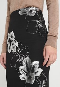 Květinová tužková sukně z černé látky s bílým květinovým potiskem, která se pyšní hladkým povrchem a přiléhavým střihem. V kombinaci s béžovým tričkem s dlouhým rukávem.