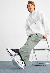Bílá Nike bunda, olivově zelené cargo kalhoty a bílo-červeno-černé tenisky s detailními prvky z síťoviny a gumy; sedící na chromové židli na koberci.