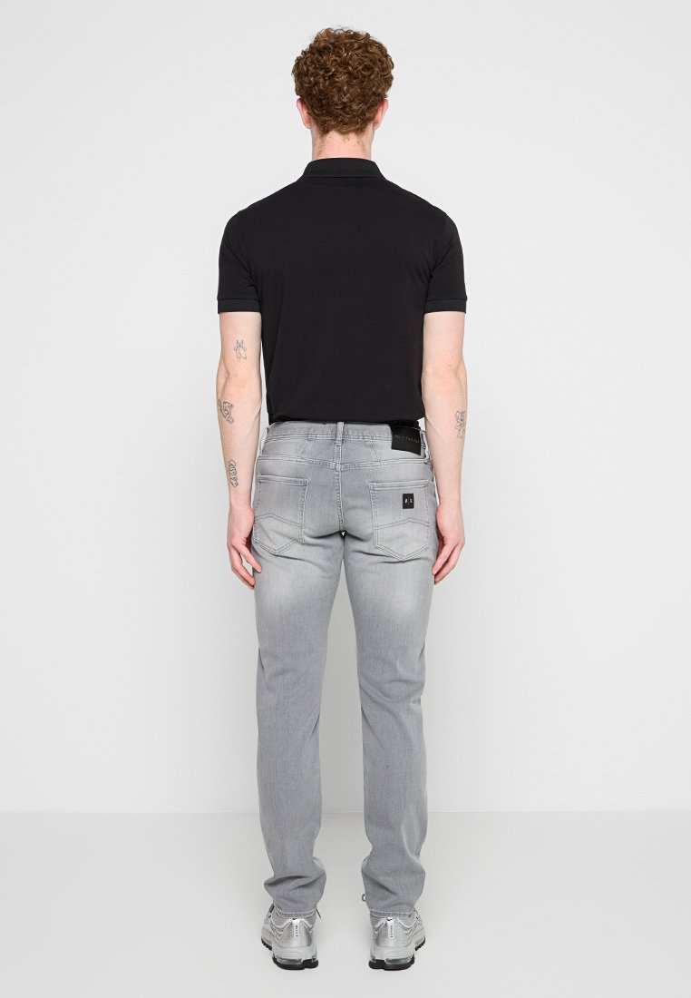 Mężczyzna z kręconymi włosami, ubrany w czarną koszulę polo, jasnoszare dżinsy i srebrne trampki, stoi przodem do gładkiej białej ściany.