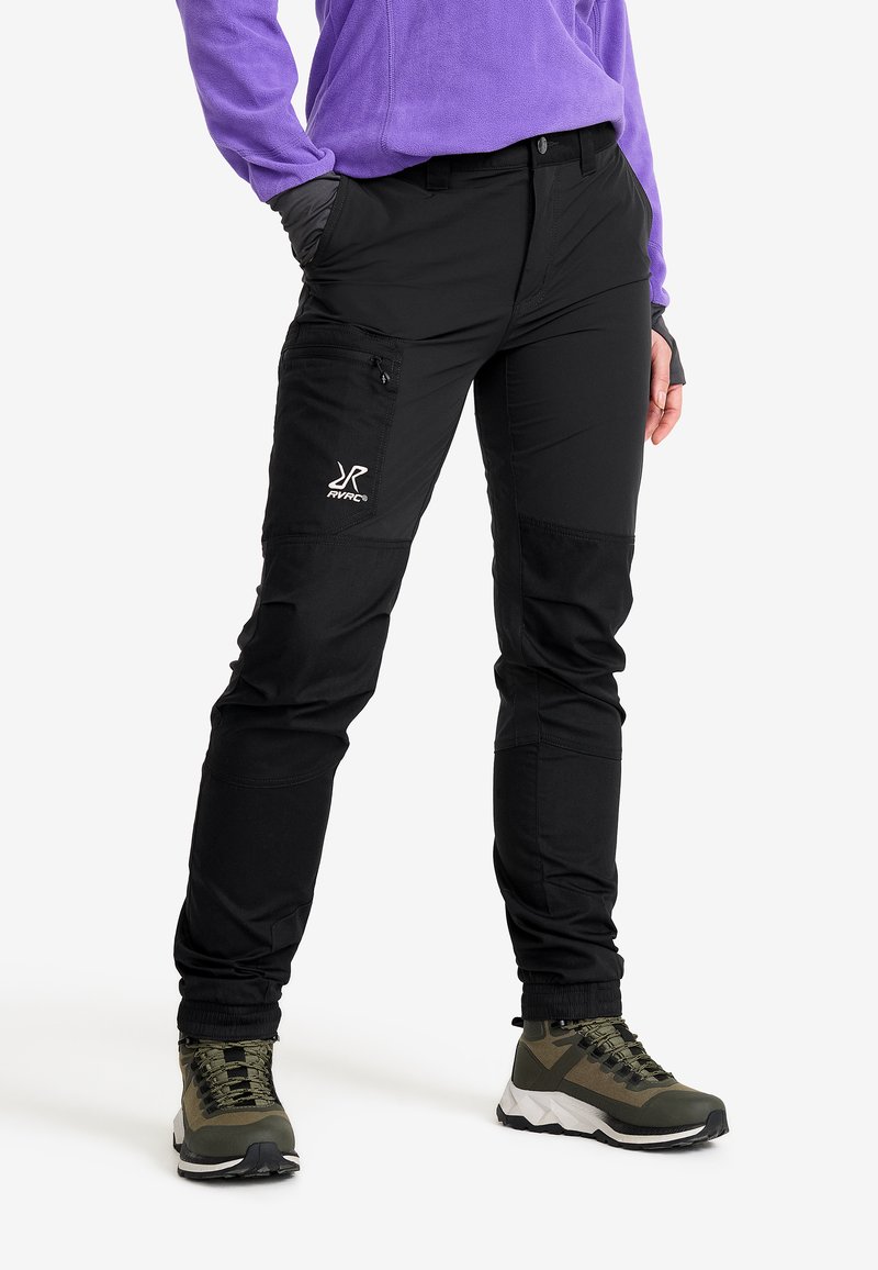 Person med svarta cargobyxor med dragkedjeficka och svart-gröna vandringsskor, i kombination med en lila långärmad topp, står med händerna i fickorna.