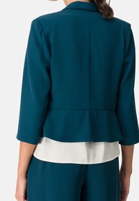 Zielony marynarka peplum z rękawami do łokci, z wyraźną konstrukcją i gładką fakturą, noszona na białej bluzce.