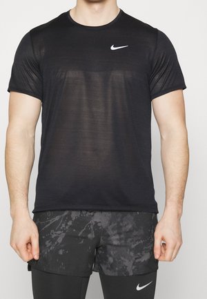 Mand iført en sort Nike kortærmet trøje og sorte mønstrede shorts over sorte leggings, stående mod en ensfarvet baggrund.