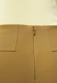 Sukně v tan barvě s hladkou texturou, která má dvě přední kapsy a středový zip. Horní část je vyrobena z jasného textilu.