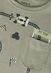 Ljusgrön bomullst-shirt med grafik av en racerbilsbild, däckillustrationer, racingflaggor och en "PIT STOP"-tagg på fickområdet.