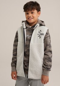 Gilet en sherpa blanc avec fermeture éclair à l'avant, porté par-dessus un sweat à capuche camo gris. Présente des lettres et des accents noirs, avec une finition douce et texturée.