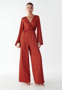 Raudonas apklijuojamo stiliaus šuoliai su ilgomis rankovėmis, giliais V formos iškirpėmis ir plačiais keliais. Lygus, lengvas audinys su subtiliu blizgesiu.