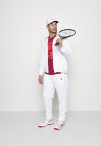 Chaqueta y pantalones deportivos blancos, camiseta roja con diseño geométrico, gorra blanca y zapatos con detalles en rosa. Sosteniendo una raqueta de tenis.