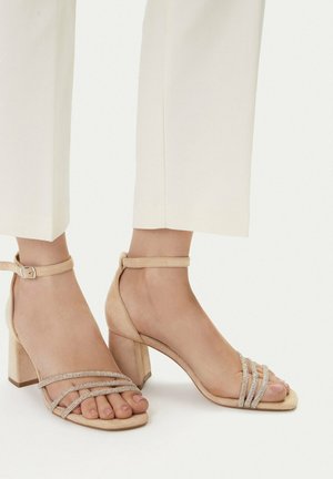 Stopy w beżowych zamszowych sandałach na blokowym obcasie, z trzema cienkimi paskami ozdobionymi kryształkami i paskami wokół kostki, zestawione z białymi skróconymi spodniami.