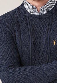 Granatowy sweter wełniany z teksturowanymi wzorami i wzorem w diamenty. Akcentowany małym haftowanym emblematem. Nosi się na koszuli w kratę.