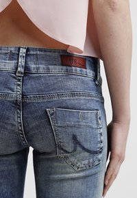 Close-up of a person wearing faded blue jeans with a leather "LTB" label and a light pink top cropped above the waist.
