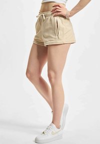 Beżowe szorty z elastycznym pasem i sznurkiem, z dwoma zapinanymi na zamek bocznymi kieszeniami oraz miękką teksturą, w połączeniu z białymi trampkami.