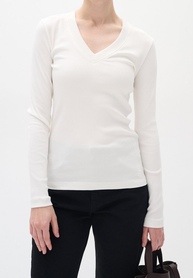 Femme portant un haut blanc cintré à manches longues et col en V, ainsi qu'un pantalon noir, tenant un sac à main marron foncé à côté d'elle.