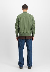 Groene bomberjack met ribbelkraag en -manchetten, zachte stof, iets gewatteerde schouders, gedragen over een bruine blouse en blauwe spijkerbroek.