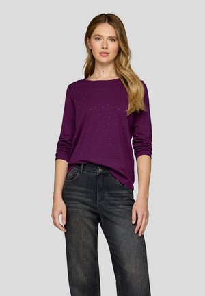 Lila Langarmshirt mit dezenten Nietenverzierungen, kombiniert mit dunklen Denim-Jeans. Die Textur ist glatt, mit lockerem Schnitt.