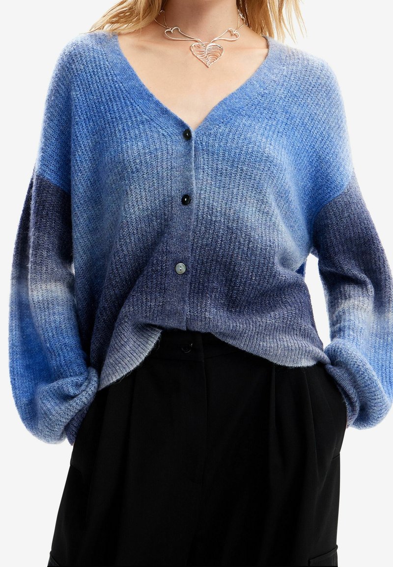 Blå gradient kofta, tillverkad av mjukt stickat tyg. Har en V-ringning, långa ärmar och knappar framtill. Texturerad med olika nyanser av blått.