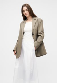 Νεαρή γυναίκα με μπεζ υπερμέγεθες σακάκι πάνω από λευκό αμάνικο τοπ και μακριά λευκή φούστα, στέκεται με το ένα χέρι στη τσέπη.