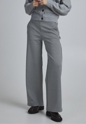 Persoon draagt een grijze cardigan met knopen, zwart-witte pied-de-poule brede broek en donkerbruine platformschoenen, staand tegen een effen achtergrond.