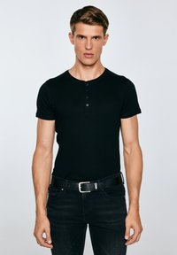 Jonge man staand, gekleed in een strak zwart henley-shirt, zwarte jeans en een zwarte riem, tegen een effen lichtgrijze achtergrond.