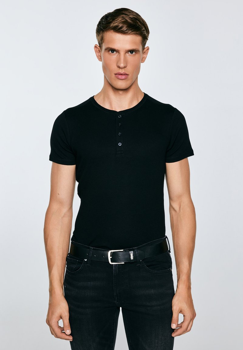 Jonge man staand, gekleed in een strak zwart henley-shirt, zwarte jeans en een zwarte riem, tegen een effen lichtgrijze achtergrond.