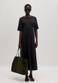 Zwart, knielang t-shirt jurkje met korte mouwen. Model houdt een donkergroene suède tote bag vast, draag oversized zwarte zonnebril en slippers.