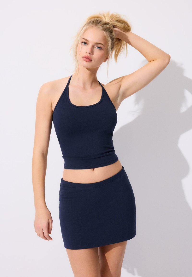 Femme blonde en crop top halter bleu marine et mini-jupe posant avec une main touchant ses cheveux sur un fond clair uni.