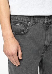 Jean en denim gris coupe droite, doté d'un design classique à cinq poches, coutures visibles et fermeture à bouton. Un bracelet argenté est porté au poignet.