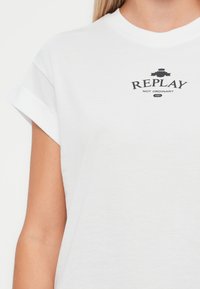 Balta kokvilnas T-kreklu ar sarullētām piedurknēm, kurā centrā ir melns dr printing logo ar uzrakstu "REPLAY NOT ORDINARY" virs gada "1981."